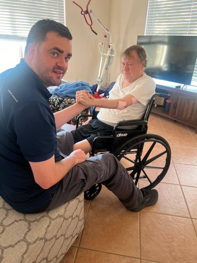 Man sitting on a couch assisting another man in a wheelchair inside a room with tiled flooring.