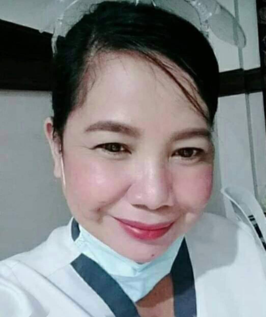 Smiling woman in a uniform, wearing a light blue surgical mask under her chin.