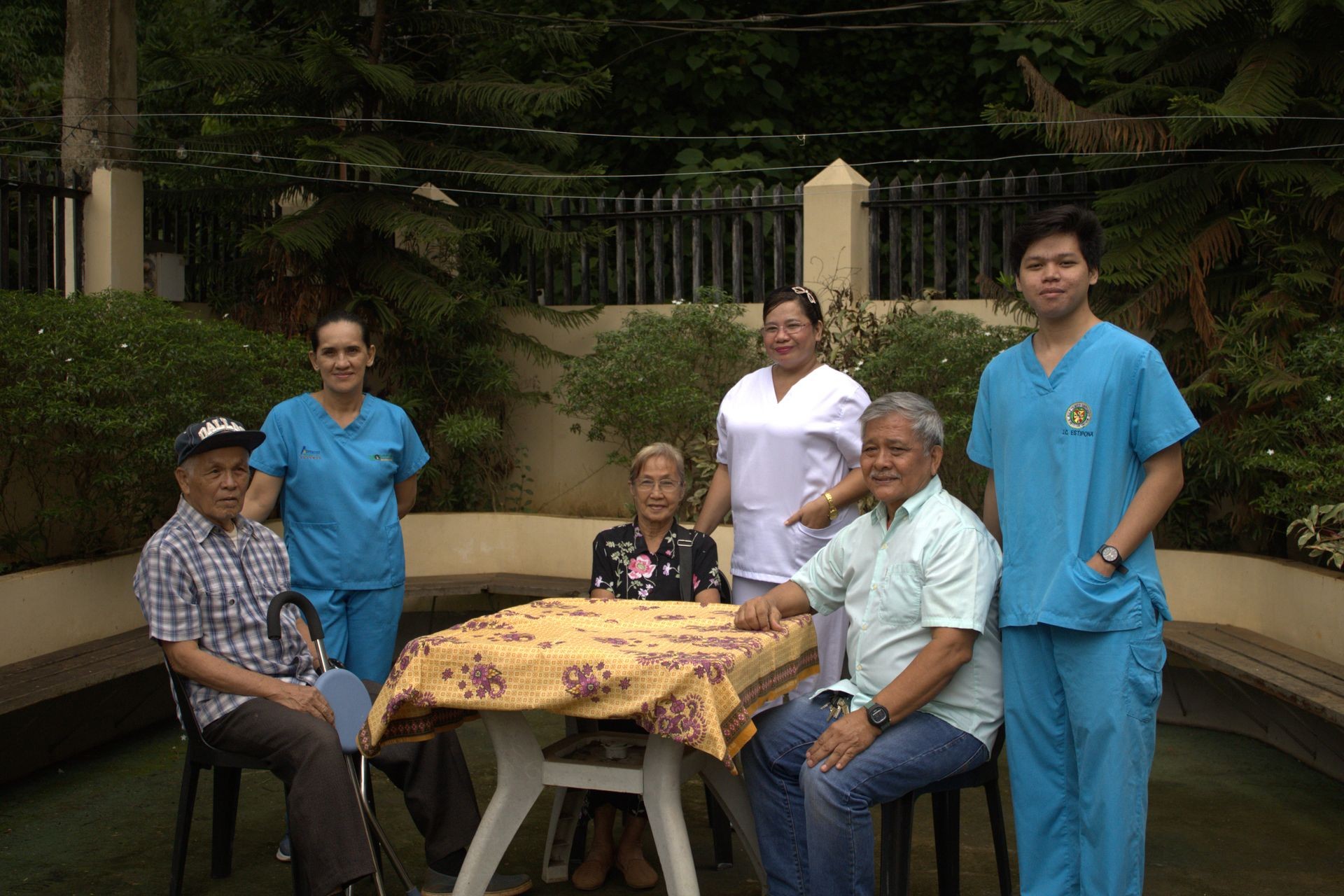 Group of elderly people and healthcare workers gathered around a table in a garden setting.