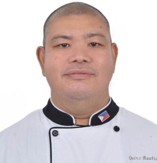 Chef wearing a white coat with black trim and a small flag emblem.